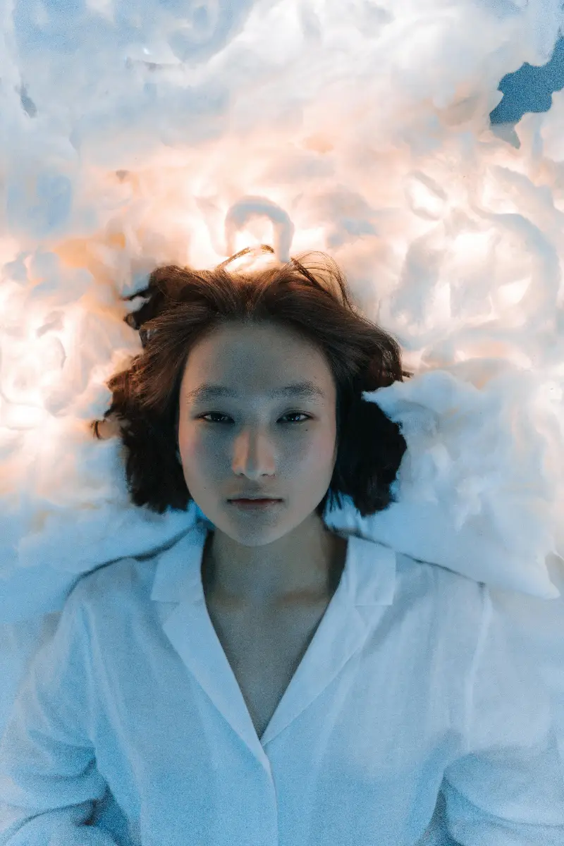 Serene portrait of an Asian woman lying on fluffy white fabric under soft lighting.